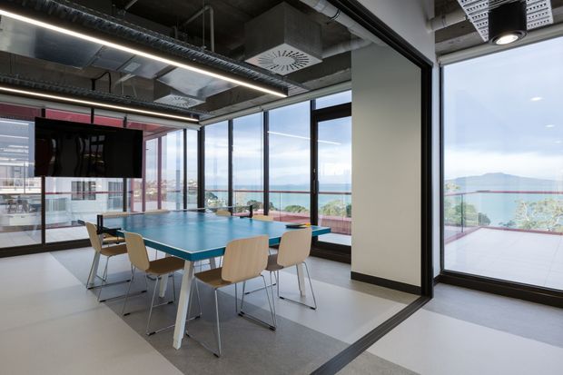 A bright modern commercial meeting and recreation space with a blue table-tennis table surrounded by chairs, a wall-mounted screen, floor-to-ceiling glazing, and an adjacent outdoor terrace with expansive water and mountain views. The exposed ceiling services, ducts, linear lighting, and black-framed glazing create an industrial-contemporary office aesthetic.
