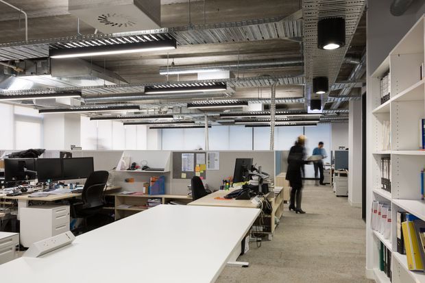 Open-plan contemporary office interior with rows of workstations, computers, office chairs, shelving, filing units, and staff moving through the space. The exposed services ceiling features metal cable trays, ducts, conduits, suspended linear fluorescent lighting, and visible concrete surfaces, giving the workplace an industrial character. White desks, partitions, shelving, and grey flooring create a bright, functional commercial environment.