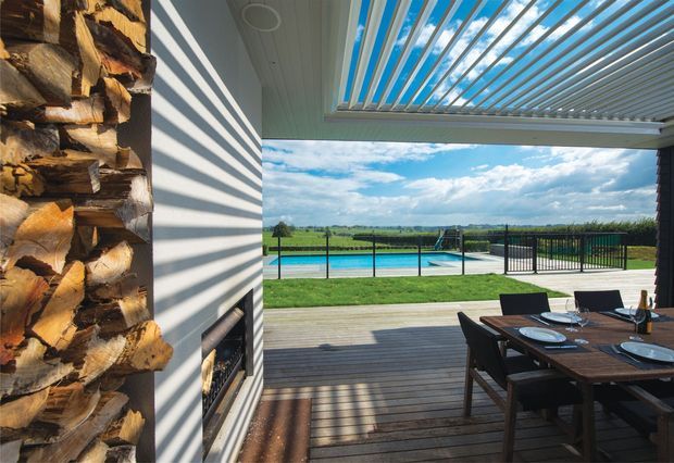 Covered outdoor dining and entertaining area opening onto a timber deck, with a louvred roof, built-in fireplace, timber-clad feature wall, dining table, swimming pool and expansive rural landscape beyond.