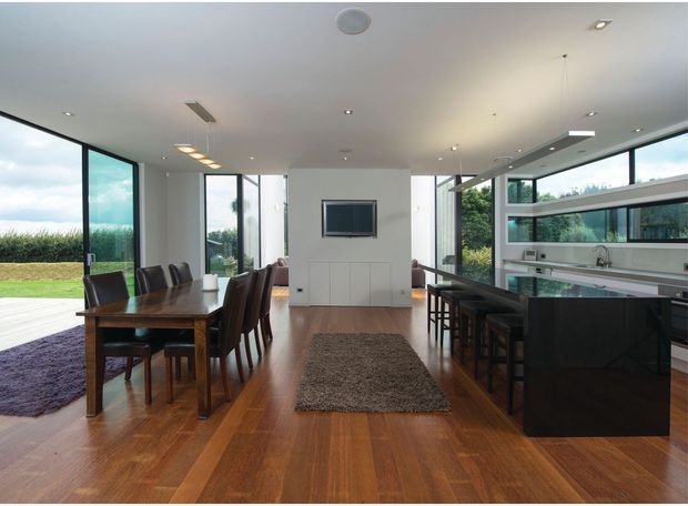 Open-plan contemporary dining and kitchen interior with warm timber flooring, a dark timber dining table and upholstered chairs, a central wall-mounted television, and a black kitchen island with bar seating. Large floor-to-ceiling windows and sliding glass doors provide abundant natural light and views of greenery and a deck. The room has a clean, spacious fit-out with recessed and linear ceiling lighting.