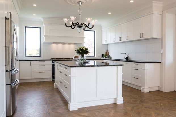 Bright, spacious traditional kitchen with extensive white shaker-style cabinetry, a large central island, black stone benchtops, stainless-steel appliances, and a warm herringbone timber floor. A decorative black chandelier, ornate ceiling medallion, substantial cornices, and classical trim add a refined heritage-inspired character.