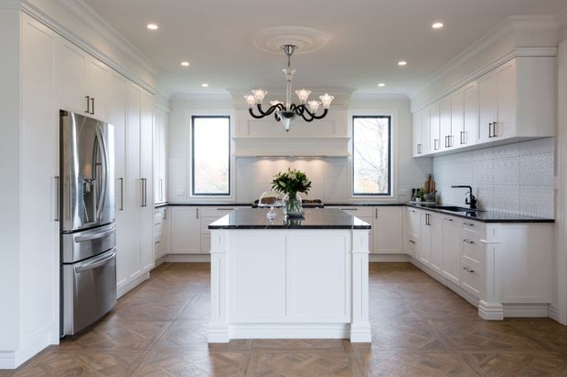 Bright, spacious traditional-style kitchen with extensive white raised-panel cabinetry, a central island with a dark stone benchtop, stainless-steel refrigerator, patterned light backsplash, integrated rangehood surround, and a decorative black chandelier. Herringbone timber flooring and tall windows add warmth and symmetry.