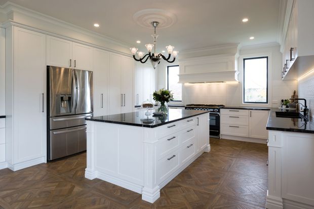 A bright, spacious traditional-style kitchen with extensive white shaker cabinetry, a large central island with a dark polished stone benchtop, stainless-steel French-door refrigerator, integrated rangehood and gas cooktop. The room features ornate ceiling cornices and a decorative ceiling medallion, recessed lighting, a black-and-crystal chandelier, and a warm herringbone timber floor. Tall windows provide abundant natural light.