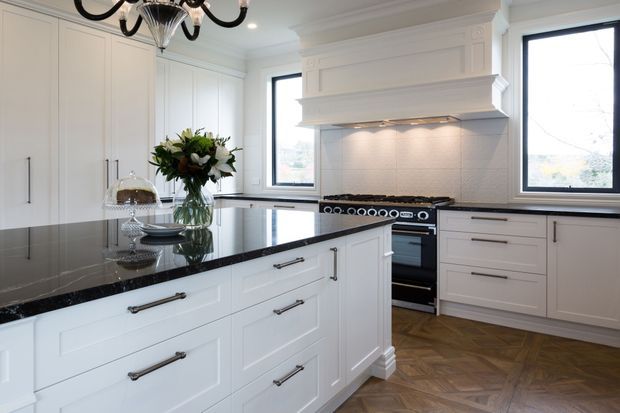 A bright, refined traditional kitchen with white Shaker-style cabinetry, a large central island, black stone benchtops, a freestanding range cooker, and a decorative white rangehood surround. Black-framed windows and a herringbone timber floor add contrast, while a chandelier and floral arrangement provide classic finishing details.