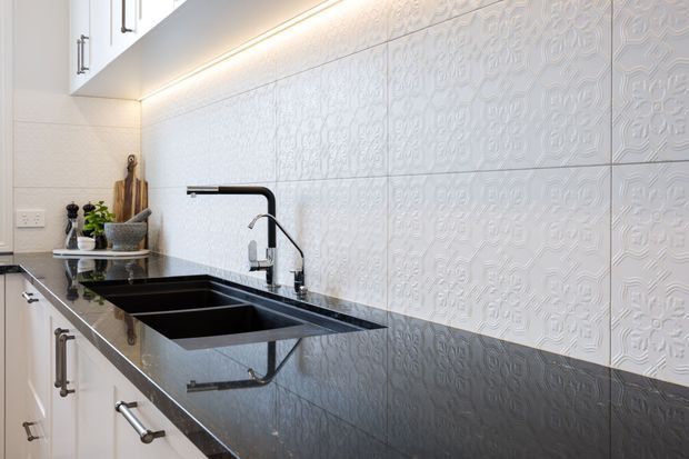 Close-up view of a bright contemporary kitchen, featuring white shaker-style cabinetry with dark metal pulls, a large black undermount double-bowl sink, black gooseneck tapware, and a polished dark stone benchtop. The backsplash is composed of white embossed patterned tiles, highlighted by warm under-cabinet lighting.