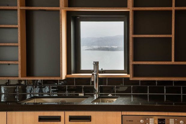 Compact modern kitchen with dark open shelving and warm timber cabinetry surrounding a central window. A stainless-steel sink and mixer sit beneath the window, with glossy dark rectangular tile splashback, timber-toned benchtop, black hardware, and integrated appliances. A hazy waterfront or lake landscape is visible through the window.