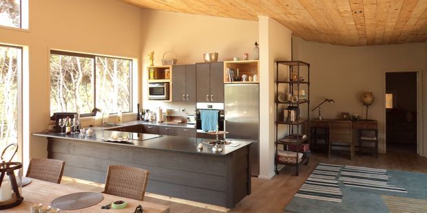 Open-plan kitchen and dining area with a large dark island, integrated cabinetry, stainless-steel refrigerator, timber ceiling, and expansive windows overlooking trees. A dining table occupies the foreground, while a styled shelving unit and console sit in the adjacent living area.