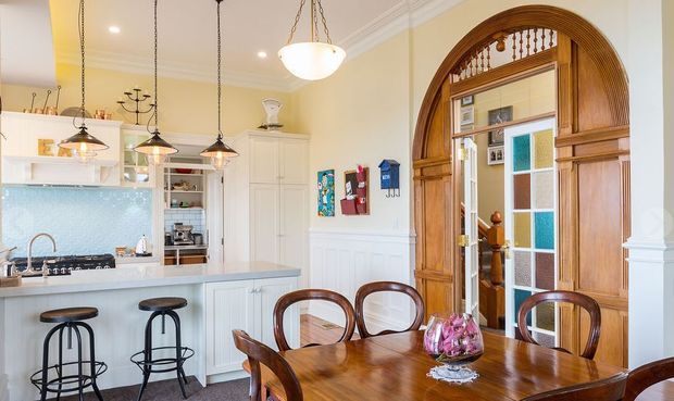 Bright, traditional-style open-plan kitchen and dining area. The kitchen features white cabinetry, a pale blue textured splashback, light benchtops, pendant lighting and a breakfast counter with stools. A polished timber dining table and chairs sit beside an ornate arched timber-framed glazed doorway with decorative fretwork and coloured glass panels.
