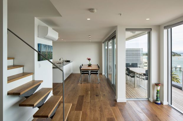 Bright contemporary open-plan interior showing a dining area beside a compact kitchen, with floating timber stair treads and a slim dark metal balustrade in the foreground. Full-height glazing and glazed doors open to a timber deck with water and urban buildings visible beyond. The room has white walls and ceiling, warm timber flooring, recessed lighting, and a clean minimalist fit-out.