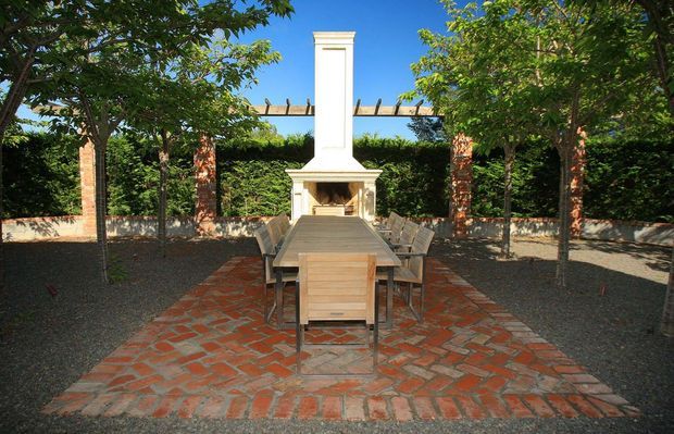 A formal outdoor dining courtyard arranged around a long timber dining table, with a white masonry fireplace and chimney at the far end. Red brick paving defines the dining area, while brick columns, a timber pergola, clipped hedging and mature trees create an enclosed garden setting.