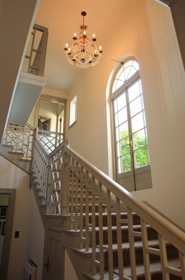 A grand interior stair hall with a multi-level staircase, painted light railings and geometric balustrades, patterned stair runner, tall arched multi-pane window, and an ornate crystal chandelier. Warm cream walls and abundant daylight create an elegant, traditional atmosphere.