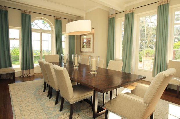 Elegant formal dining room with a large dark wood rectangular dining table surrounded by upholstered cream dining chairs. The room features tall arched and rectangular windows, layered teal and cream curtains, dark timber flooring, a patterned area rug, traditional wall mouldings, and a large contemporary drum pendant light. The space is bright, refined, and classically styled.