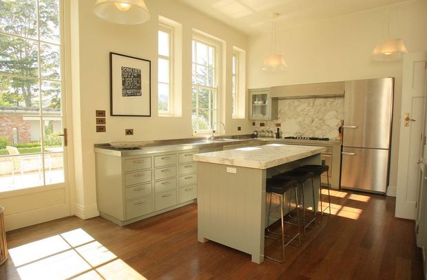 Bright, spacious traditional kitchen with tall multi-pane windows and glazed doors opening to a garden or outdoor area. The room features a large central island with a pale stone benchtop and seating, extensive light-coloured cabinetry, dark timber flooring, a stainless-steel refrigerator, pendant lights, and a marble-patterned splashback behind the cooktop.