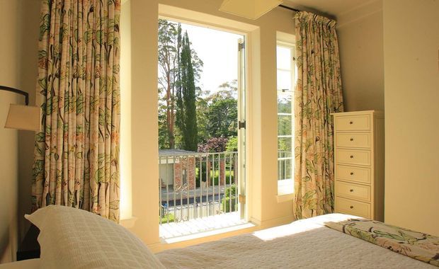 Bright traditional-style bedroom with a large glazed door opening onto a small balcony, tall patterned botanical curtains, cream walls, a pale upholstered bed, bedside lamp, and a narrow white chest of drawers. Trees and garden are visible beyond the balcony.