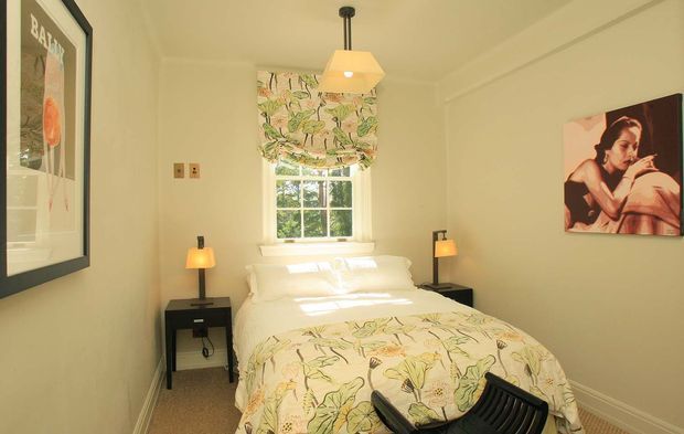 A compact, warm bedroom with a double bed dressed in a botanical floral duvet and matching Roman blind, positioned beneath a multi-pane window. The room has cream walls, carpeted flooring, small dark bedside tables with lamps, a central pendant light, framed artwork, and simple traditional detailing.