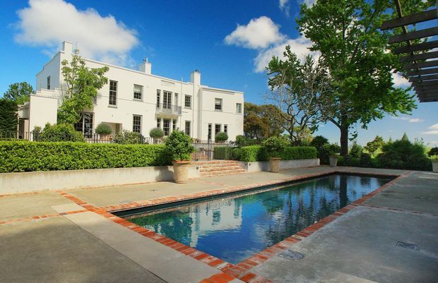 A large white multi-storey residence with a restrained classical facade, tall sash-style windows, small balconies and symmetrical detailing, viewed from a landscaped pool terrace. The rectangular swimming pool has red brick coping and sits within a broad paved courtyard bordered by clipped hedges, mature trees and a pergola-like overhead structure. Bright blue sky and reflective water create a sunny, elegant outdoor setting.