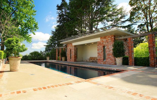 A landscaped outdoor pool terrace with a long rectangular swimming pool, pale paved deck, and a low modern pool pavilion. The pavilion features exposed red brick walls, a broad flat roof with deep overhangs, large open bays, and timber accents, surrounded by mature trees, hedges, and potted plants.