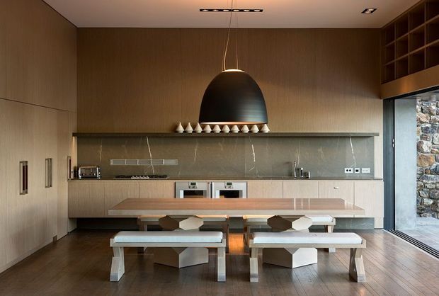 A refined open-plan kitchen and dining area with full-height warm timber cabinetry and wall panelling, a long muted stone backsplash, integrated appliances, and a broad timber island or dining counter. Contemporary pendant lighting hangs above the island, while built-in shelving and a large opening reveal a stone exterior wall. Two modern dining benches and geometric chairs occupy the foreground, with dark timber flooring throughout.