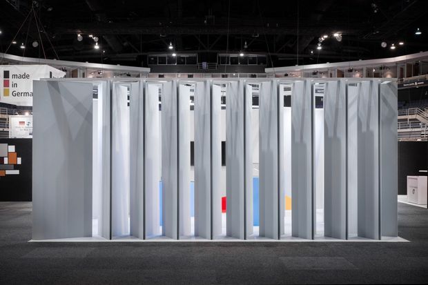 A contemporary trade-show exhibition display featuring a long row of tall, narrow, vertically folded or pleated light-grey architectural panels and screens. The installation is set within a large dark exhibition hall with overhead spotlights, surrounding booths, signage, and carpeted flooring. Small glimpses of colourful product displays are visible through the gaps between the panels.