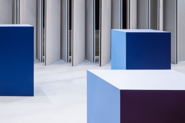Abstract contemporary architectural interior or installation featuring a white floor and a backdrop of vertical ribbed wall panels with metallic or grey framing. Three large, sharply geometric rectangular plinths or volumes in saturated blue, pale blue, white, and deep purple create a bold, high-contrast composition.