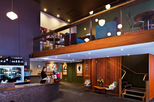 A multi-level commercial hospitality interior, likely a café, bar, or restaurant, with a curved tiled service counter in the foreground, refrigerated display cabinets, a mezzanine with timber balustrading and glass panels, and a staircase screened by vertical timber battens. The space has dark painted walls and ceilings, warm timber joinery, pendant globe lighting, and a moody, contemporary atmosphere.
