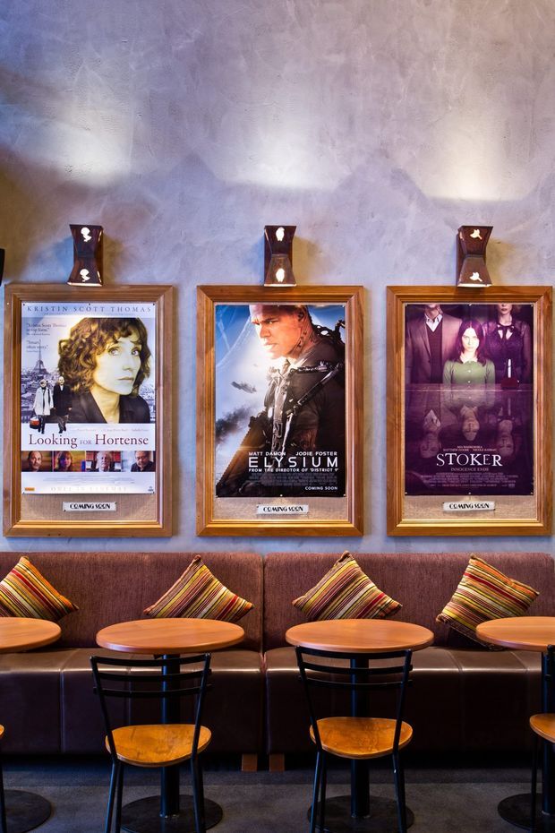 A cinema or theatre lounge with a textured lavender-grey plaster wall displaying three framed film posters in illuminated display cases. A long dark upholstered banquette runs beneath the posters, paired with small round timber tables and black metal café chairs. Striped multicolour cushions add pattern and warmth, while dark carpet and warm lighting create an intimate, theatrical atmosphere.