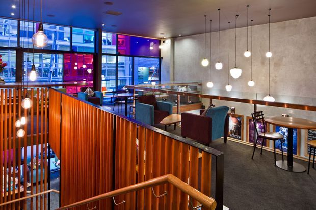 A lively multi-level hospitality interior, likely a bar or restaurant, viewed from a stairway. The space features warm vertical timber slat balustrades, dark carpet, concrete-textured walls, extensive glazing, exposed-looking services, and numerous pendant lights. Small tables and upholstered dining chairs occupy the upper level, with vivid blue, purple, and red lighting visible through the windows and lower-level openings.