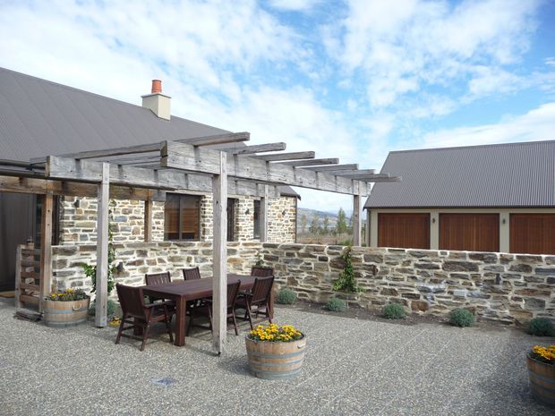 A rustic stone-clad residential courtyard or patio with a weathered timber pergola over a dark outdoor dining table and chairs. Gravel paving, raised barrel planters with yellow flowers, low stone walls, and adjacent corrugated metal-roofed buildings create a rural, traditional outdoor setting.