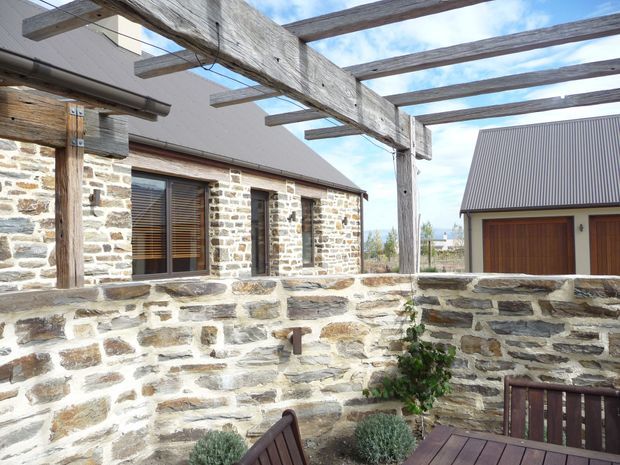 Exterior courtyard view of a rustic stone-clad residence, featuring a heavy timber pergola, low stone boundary walls, timber-framed glazing with horizontal blinds, and a detached garage or outbuilding with a corrugated metal pitched roof and timber doors. The setting appears bright and open, with a glimpse of distant landscape beyond the buildings.