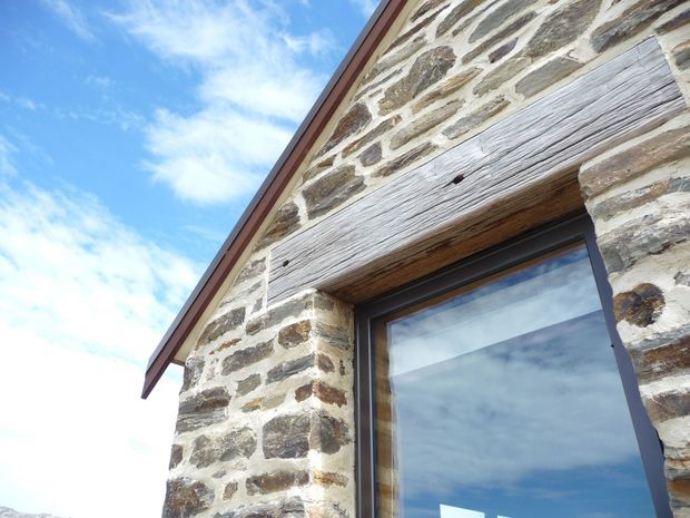 Close-up exterior view of a gabled stone building facade, showing irregular natural stone masonry with light mortar joints, a weathered timber lintel, and a dark-framed glazed opening. The roof edge and bright blue sky are visible from a low upward angle.