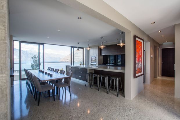 Open-plan contemporary kitchen and dining area with a long dark dining table, upholstered dining chairs, a central kitchen island with bar stools, dark cabinetry, pendant lighting, polished concrete or terrazzo-style flooring, and expansive floor-to-ceiling glazing overlooking a mountainous waterfront landscape. A hallway and framed artwork are visible to the right.