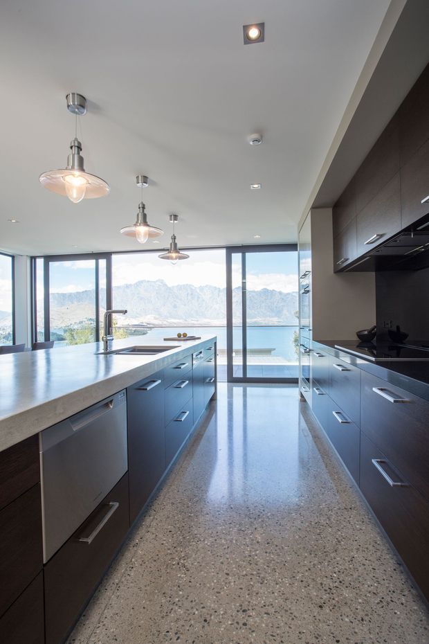 A contemporary galley-style kitchen with a large central island, dark wood cabinetry, black benchtops, stainless-steel appliances, and pendant lighting. Full-height glazing at the far end opens to a bright lakeside view with mountains beyond. The polished speckled floor and expansive windows create a bright, refined interior.