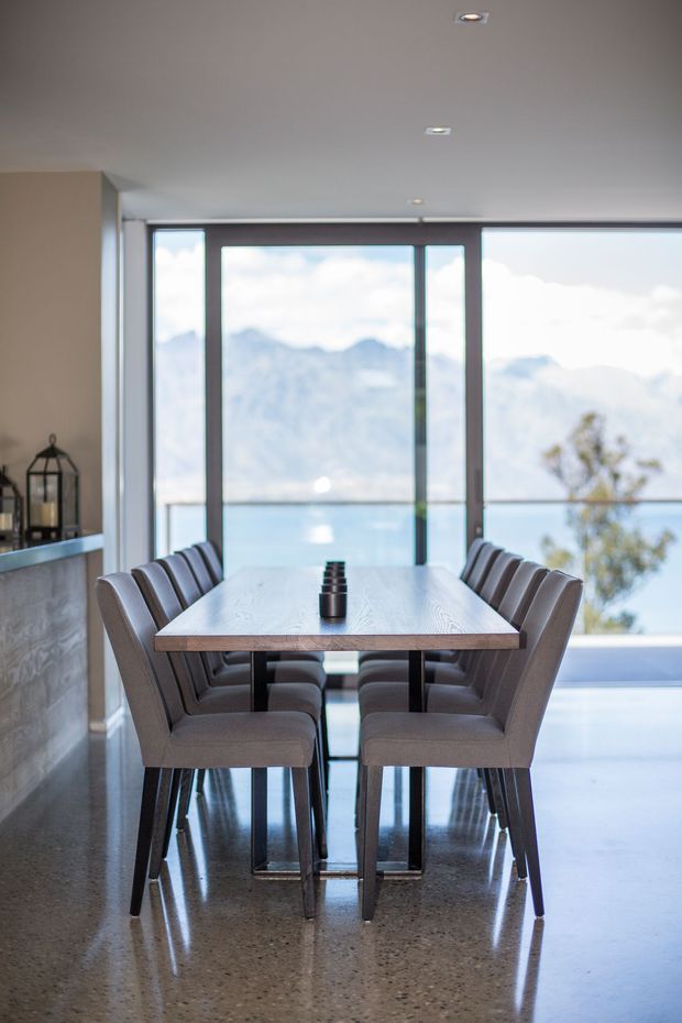 A refined dining area with a rectangular timber dining table surrounded by upholstered dining chairs. The room has a polished concrete floor, a neutral painted ceiling and walls, dark window and door frames, and expansive floor-to-ceiling glazing overlooking a blue lake and mountainous landscape. A partial kitchen or built-in counter is visible at left, with recessed ceiling lighting and a small decorative lantern.