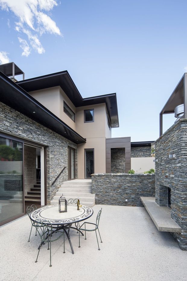 A modern multi-level residential courtyard with extensive stacked-stone walls, dark roof overhangs, smooth rendered upper walls, and contemporary glazing. The paved courtyard includes a small ornate metal outdoor dining table and chairs, built-in stone bench seating, steps, and landscaped pockets. The composition emphasizes the enclosed outdoor entertaining space and layered architectural forms under a clear blue sky.