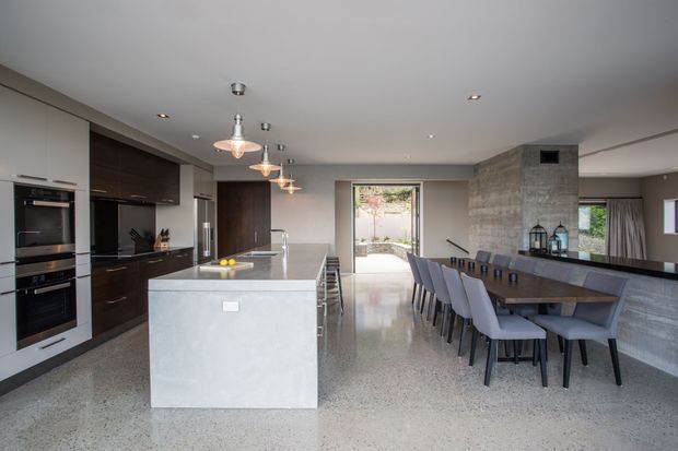 A spacious open-plan kitchen and dining area with a contemporary, minimalist fit-out. The kitchen features dark wood-toned full-height cabinetry, integrated appliances, a large pale stone island with seating, and pendant lights. A long dark dining table with upholstered grey chairs sits beside a textured grey concrete feature wall and built-in dark cabinetry. Polished terrazzo flooring, recessed ceiling lights, and large openings create a bright, refined interior.