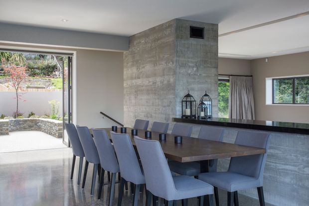 Contemporary open-plan dining area with a long dark timber dining table and upholstered chairs, positioned beside a polished concrete feature wall and built-in concrete-faced fireplace or structural volume. Large sliding glazing opens to a landscaped courtyard, while neutral walls, recessed lighting, and generous windows create a refined indoor-outdoor connection.