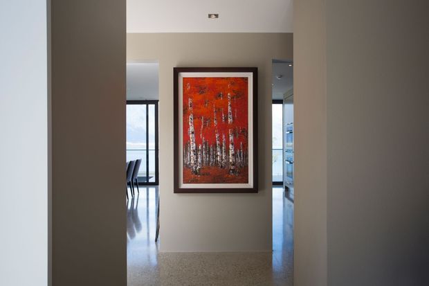 A narrow interior hallway or entry passage with warm taupe walls, a polished light terrazzo or concrete floor, and a large framed red-orange artwork as the focal point. Openings on either side reveal a dining area with floor-to-ceiling glazing and a bright adjoining room. The space is minimalist and contemporary, with simple recessed ceiling lighting and clean architectural lines.