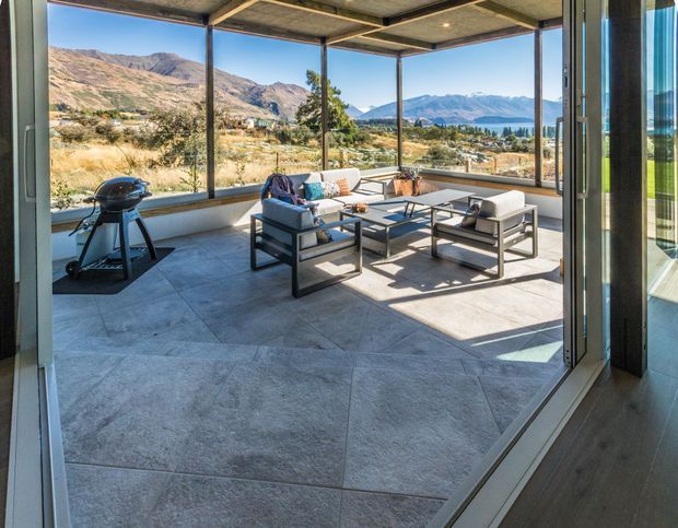 A bright, enclosed outdoor living terrace with floor-to-ceiling glazing, a large tiled floor, contemporary lounge seating, and a barbecue. The space opens to expansive mountain and lakeside views, with slim dark structural framing and a flat roof creating a modern pavilion-like form.