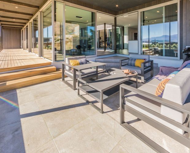 A bright, spacious contemporary outdoor lounge terrace adjoining a glazed modern home. The covered patio has large pale stone-look floor tiles, timber steps and decking, dark ceiling lining, extensive floor-to-ceiling glazing, and grey outdoor sofas and armchairs arranged around low rectangular coffee tables. Mountainous landscape is visible through the glass, with strong sunlight and subtle rainbow lens flare.