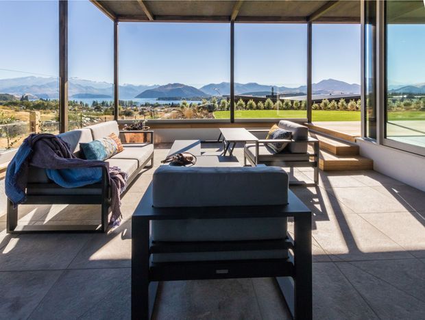 A contemporary sunroom or living area with expansive floor-to-ceiling glazing overlooking a landscaped property, lake, and mountain range. The room features a dark tiled floor, exposed timber ceiling structure, streamlined dark-framed windows, low modern sofas and armchairs, a coffee table, and stepped built-in seating along the window wall. Bright natural light and strong indoor-outdoor visual connection dominate the space.
