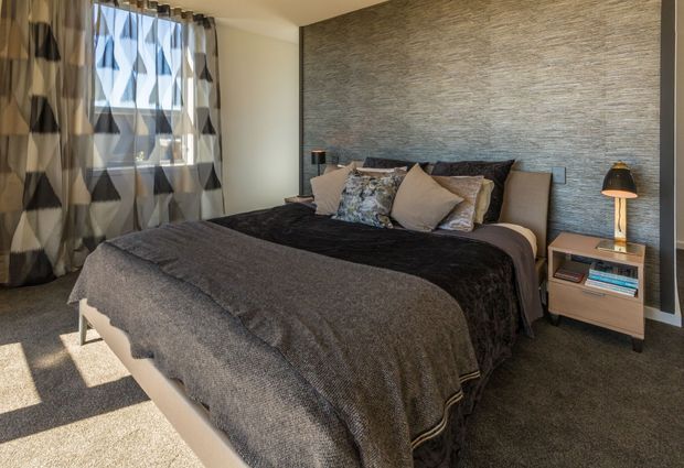 A contemporary bedroom with a large upholstered bed dressed in layered charcoal and taupe bedding, patterned and neutral cushions, and matching bedside tables with lamps. A textured grey feature wall anchors the bed, while geometric patterned curtains frame the window and warm daylight enters from the left. The room has a calm, refined, tonal palette and soft furnishings.