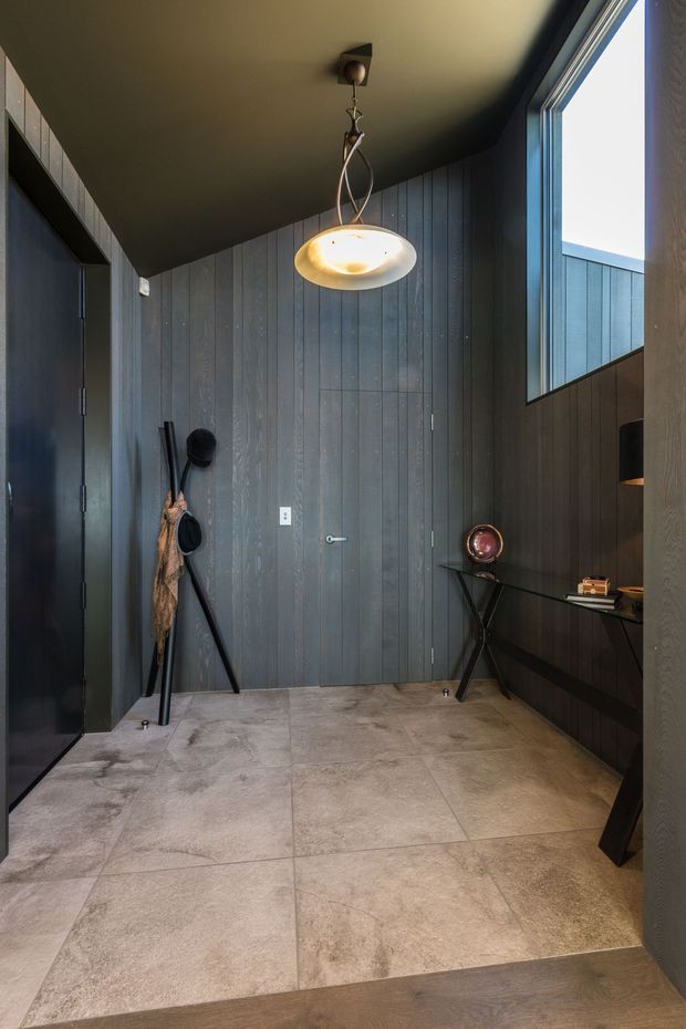 A dark, refined entryway with vertical charcoal timber wall panelling, a matching dark ceiling and door, large stone-look floor tiles, and a sculptural pendant light. A narrow console shelf with decorative objects sits beneath a tall window, while umbrellas and walking sticks lean against the wall. The space has strong vertical lines, warm accent lighting, and a contemporary, moody character.