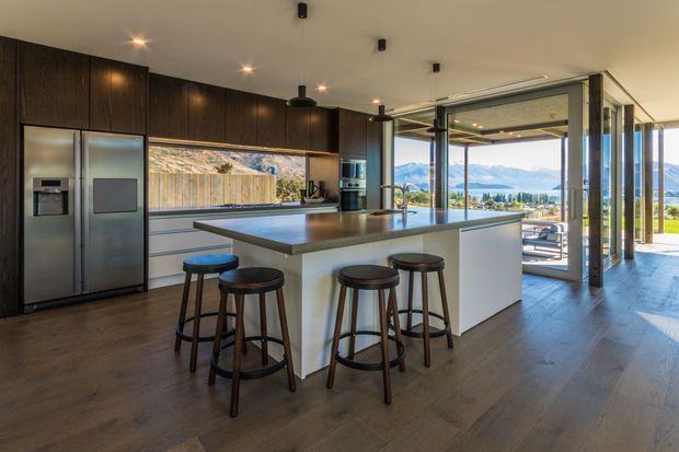 Contemporary open-plan kitchen dominated by a large island with seating for four. Dark timber cabinetry, integrated stainless-steel appliances, a pale island base and dark benchtop create a refined contrast. The kitchen opens through extensive glazing and large sliding doors to a covered outdoor area, with mountains visible beyond.