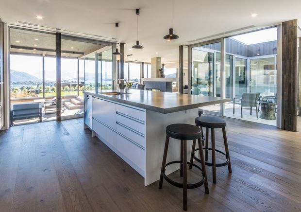 Bright, spacious contemporary kitchen with a large white island, dark stone benchtop and integrated seating. Full-height glazed doors and windows open onto a furnished outdoor terrace and courtyard, with distant mountain views visible beyond. The room features dark timber flooring, exposed dark-framed glazing, minimalist cabinetry and black pendant lights.