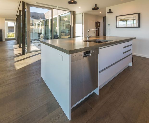 A bright contemporary kitchen centred on a large white island with a dark grey stone benchtop, integrated sink and stainless-steel dishwasher. The room features timber flooring, white walls, black pendant lights, extensive glazed doors, and views into a courtyard. Fixed cabinetry and clean-lined detailing create a modern, open-plan feel.