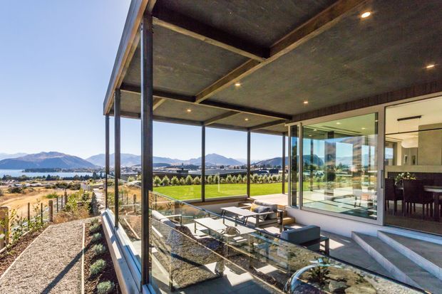 A contemporary outdoor terrace and glazed living area overlooking landscaped grounds, a lake, and distant mountains. The terrace has dark metal posts, extensive glass balustrades and glazing, a concrete soffit with exposed timber framing, recessed downlights, outdoor lounge furniture, and a dining setting. The adjacent interior appears to contain a kitchen or dining area with dark cabinetry and a marble or stone island.