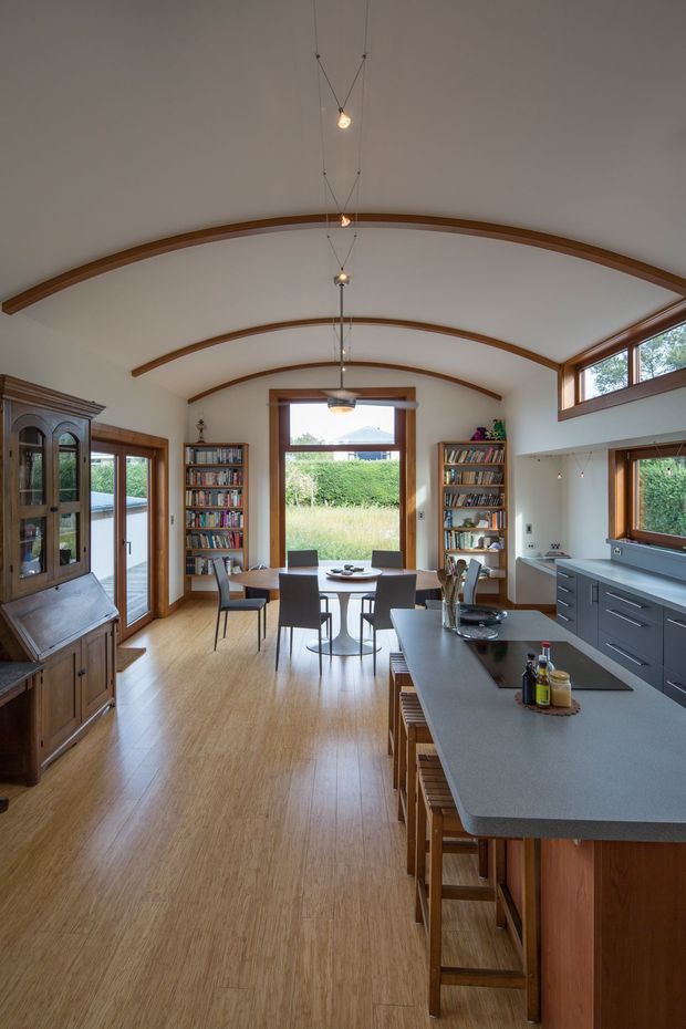 A warm, open-plan kitchen and dining space with a long island, dark grey benchtop, timber cabinetry and stools, adjacent dining table, bookshelves, and a traditional timber display cabinet. The room features distinctive curved timber ceiling beams, white plastered ceiling and walls, timber-framed windows and doors, and abundant natural light overlooking greenery. Pendant lights hang along the length of the vaulted ceiling.