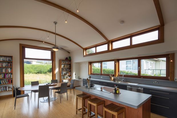 A bright open-plan kitchen and dining area with a long central island, dark built-in cabinetry, integrated appliances, timber-framed windows, and a dining table beside a wall of bookshelves. The room features a distinctive curved, timber-trimmed vaulted ceiling with exposed arch-like ribs and high clerestory glazing. Warm timber flooring, cabinetry accents, and abundant natural light create a refined, welcoming atmosphere.