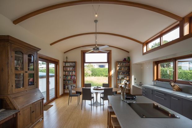 A bright open-plan kitchen and dining area with a curved vaulted ceiling and exposed timber beams. Warm timber flooring, timber-framed glazing and doors, built-in bookshelves, and a large dark kitchen island with integrated cooktop define the space. A traditional timber display cabinet and dining table add character, while clerestory windows bring in natural light and views of greenery.