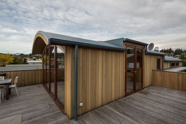 Exterior view of a compact contemporary rooftop dwelling or pavilion with warm vertical timber cladding, dark metal roofing and gutters, extensive glazing, and a large weathered timber terrace. The curved vaulted roof section creates a distinctive covered outdoor area, while a suburban landscape is visible beyond the roofline.